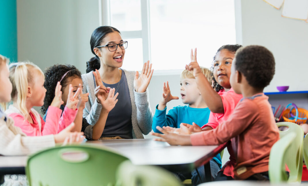 A teacher sits at a table with a diverse group of young children, engaging in a lively activity.