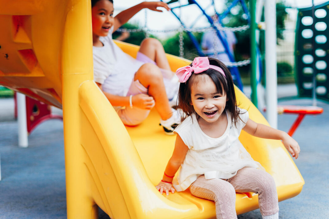 Two children are playing on a yellow slide at a playground. The child in front is smiling and sitting at the bottom of the slide, while the other is sliding down behind them.